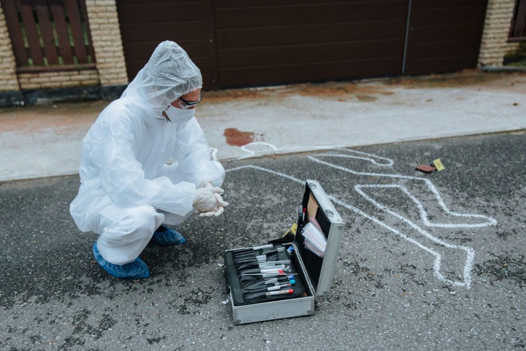 Kriminaltechniker in Schutzkleidung untersucht im Freien einen Tatort mit einem Spurensicherungskoffer.