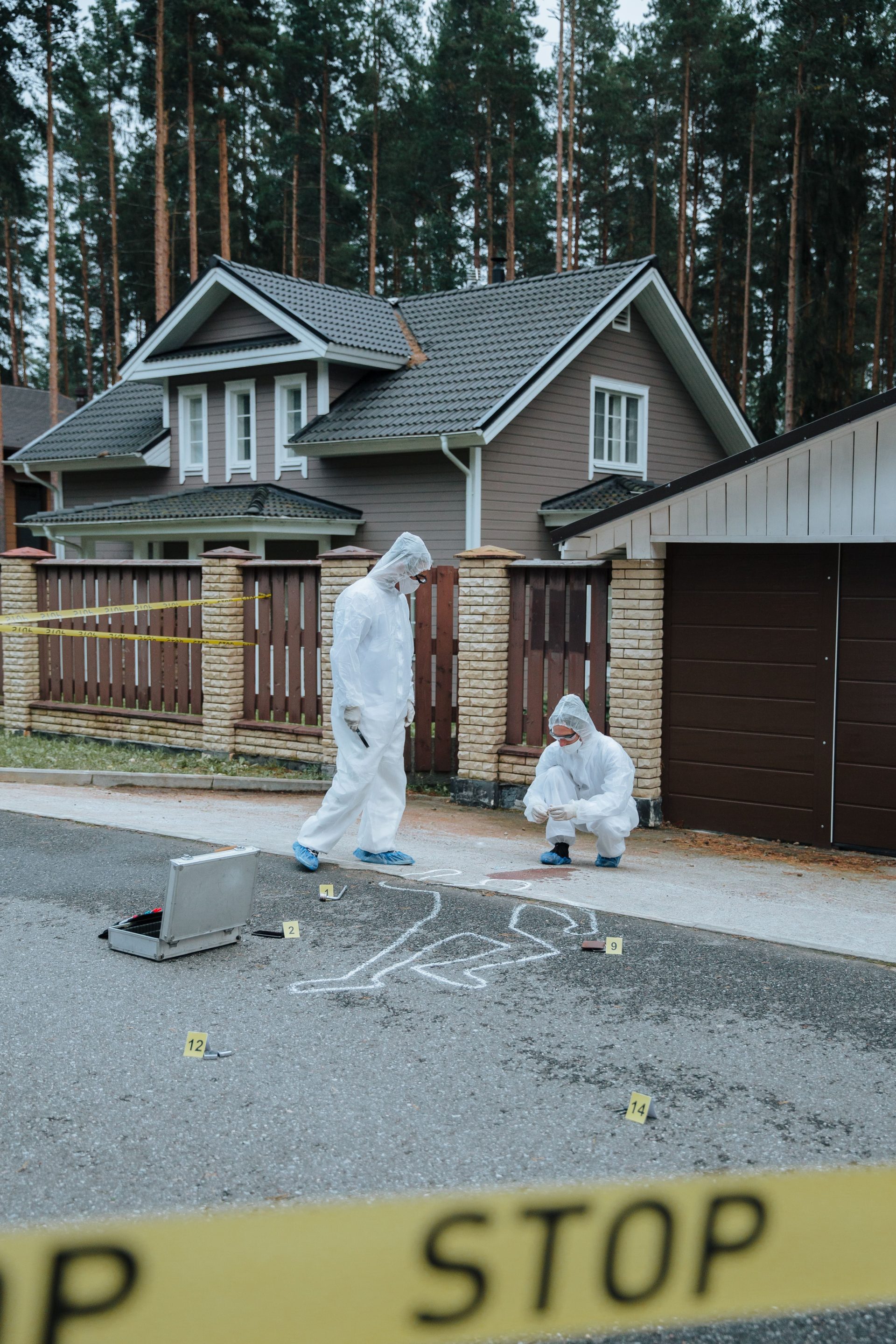 Zwei Ermittler in Schutzanzügen untersuchen einen Tatort vor einem Haus.